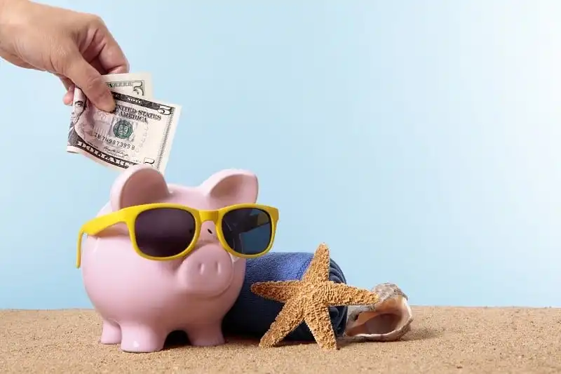Saving for beach vacation or retirement, with pink piggy bank and sunglasses. Studio shot with plain blue background. Sharp focus on the five dollar bill. Space for copy. Warm color and directional lighting are intentional.